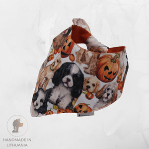 A triangular bandana-style pet bib with a white background printed with cartoon dogs and orange jack-o'-lantern pumpkins, shown on a light gray textured background. A small tag is visible at the bottom, and a 'Handmade in Lithuania' logo is in the lower-left corner.