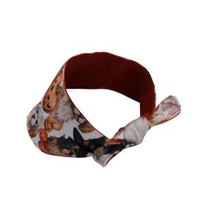 A fabric headband with a Halloween-themed print featuring pumpkins, a witch, and a dog on a white background, with a solid rust-brown inner lining and a knotted tie.
