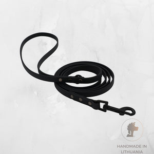 A black biothane dog leash with metal hardware, coiled on a light gray textured background. A circular logo in the bottom right corner reads 'HANDMADE IN LITHUANIA' with a lion head icon.