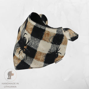 A handmade triangular pet bandana featuring a black, beige, and brown buffalo plaid pattern with black spider graphics, displayed on a light gray textured background.