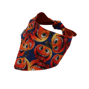 A triangular bandana-style bib with a playful print of smiling orange pumpkins on a dark blue background, featuring a solid rust-brown inner lining and strap.