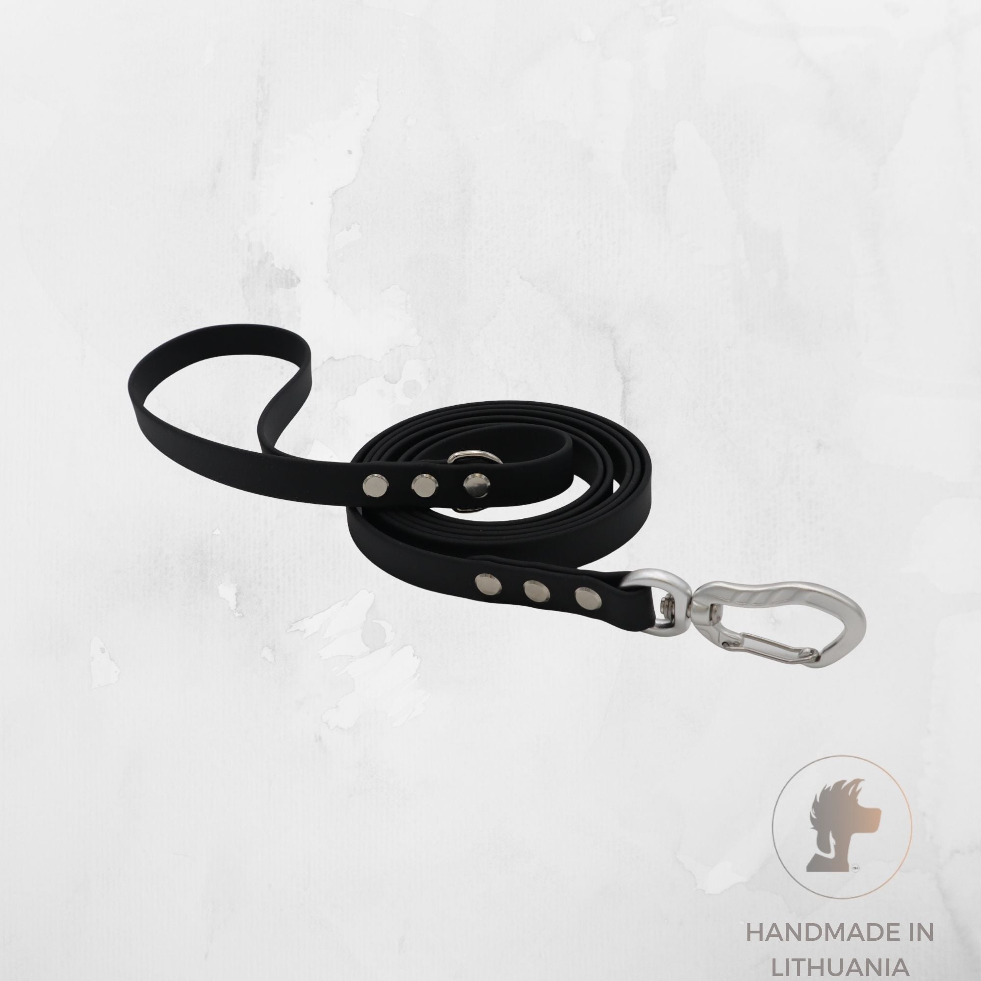 A black biothane dog leash with silver metal hardware, including a silver carabiner clip and silver rivets, displayed on a light gray textured background. A circular logo in the bottom right corner reads 'HANDMADE IN LITHUANIA' with a lion head icon.