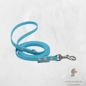 A bright blue dog leash with silver metal hardware, including a clasp and rivets, coiled on a light gray marble-textured background. A small label reads 'SVEITISMAIYAS KALE', and a circular 'HANDMADE IN LITHUANIA' logo is in the bottom right corner.