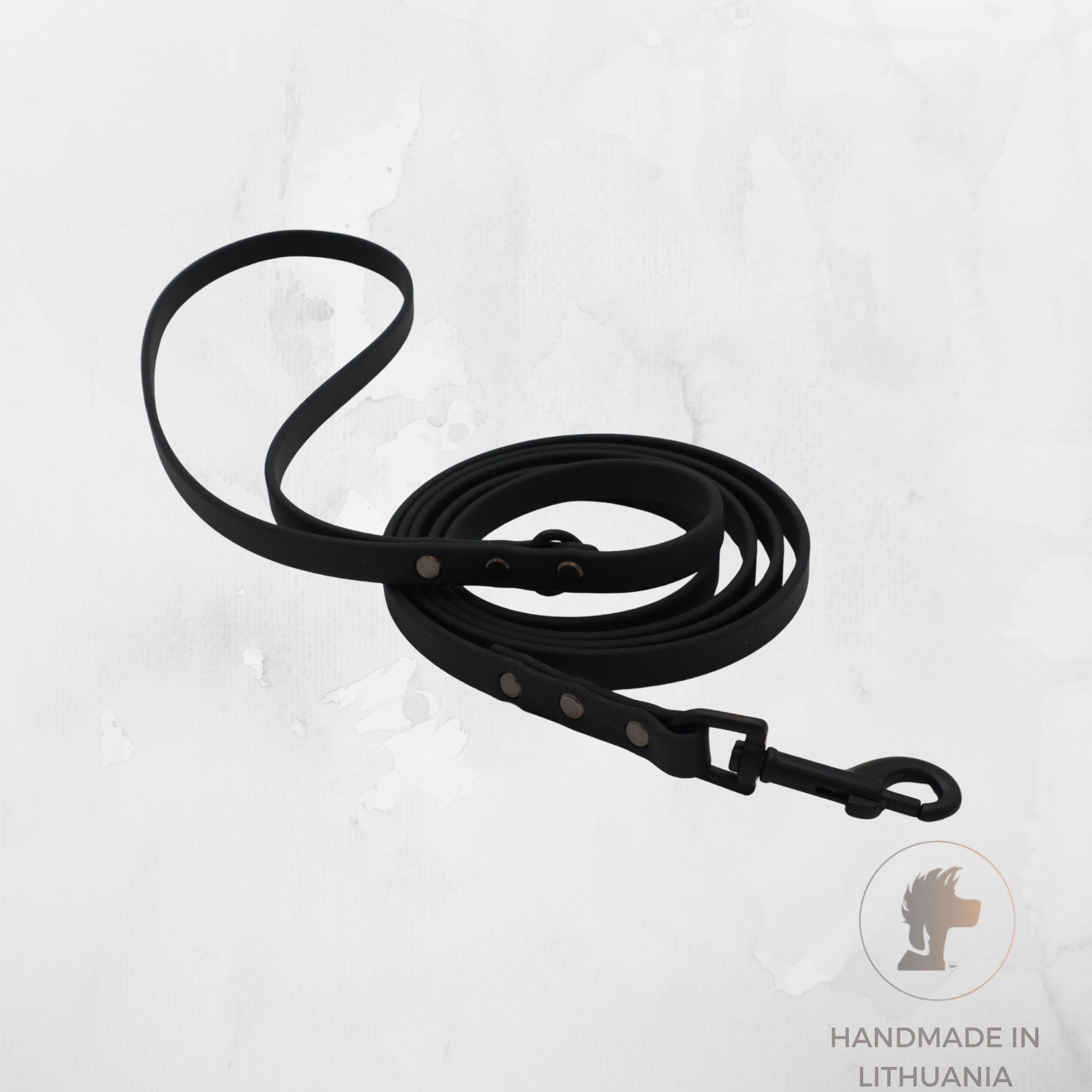 A black biothane dog leash with metal hardware, coiled on a light gray textured background. A circular logo in the bottom right corner reads 'HANDMADE IN LITHUANIA' with a lion head icon.