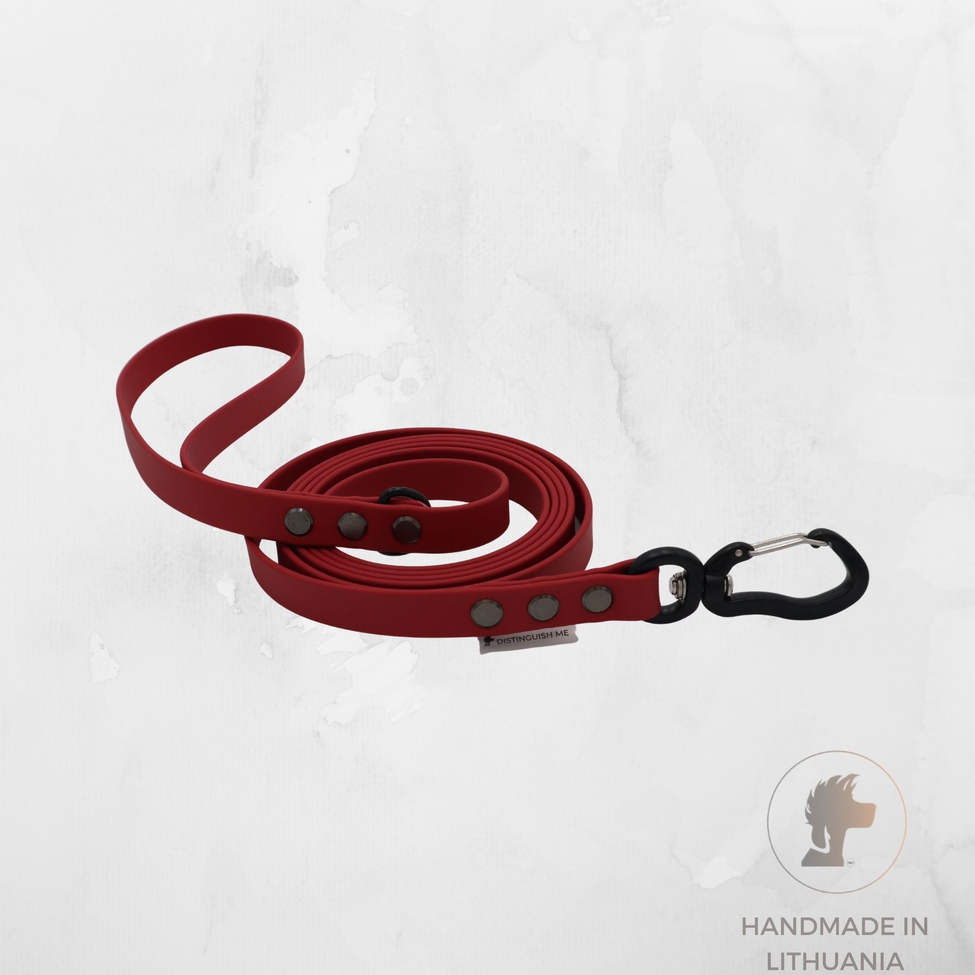 A red dog leash with a black metal carabiner clip, displayed on a light gray textured background. A circular logo in the bottom right corner reads 'HANDMADE IN LITHUANIA' with a lion head icon.