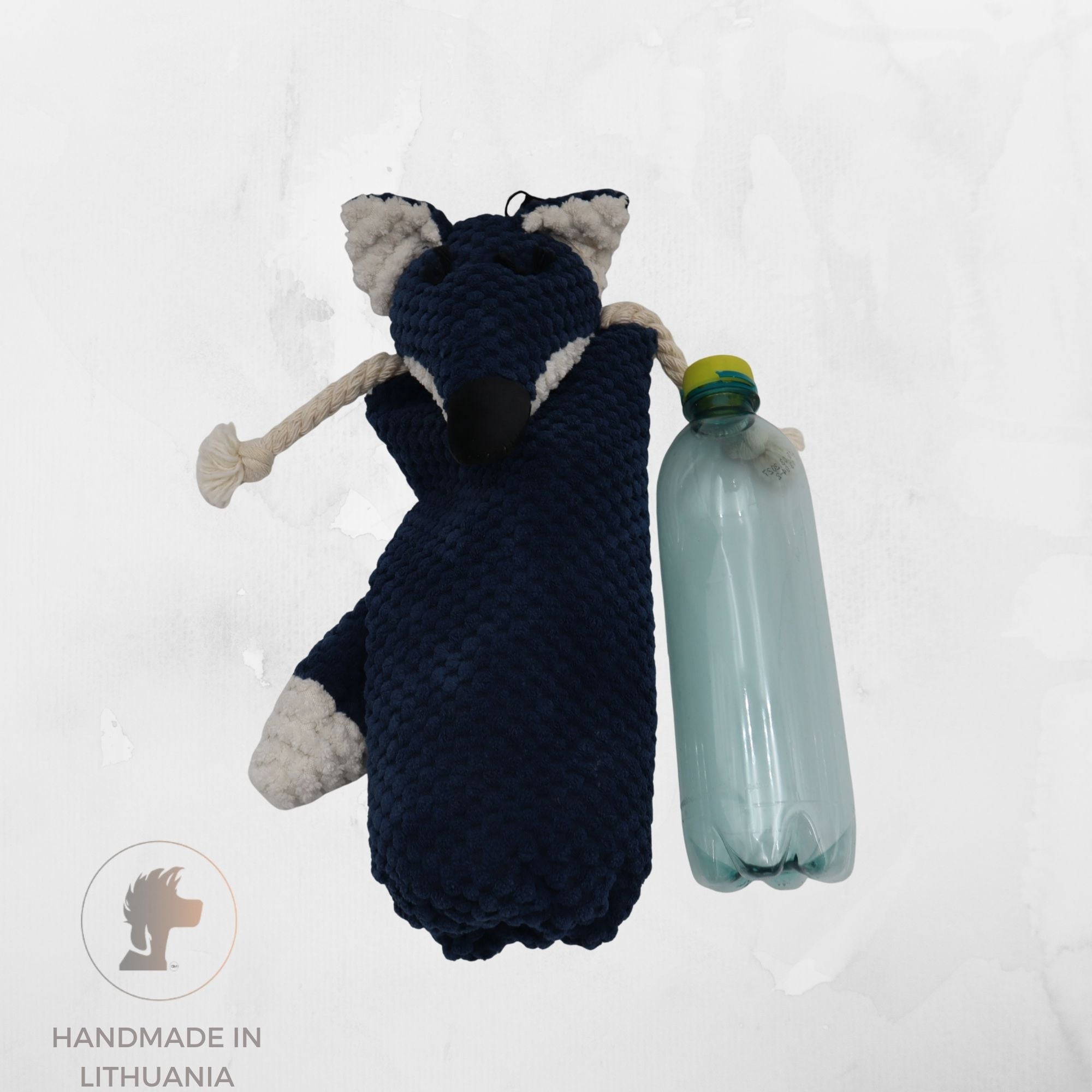 A handmade knitted fox-shaped dog toy in navy blue and white, holding a clear plastic bottle, shown against a light gray textured background with a 'Handmade in Lithuania' logo in the bottom-left corner.