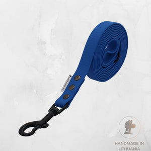A royal blue dog leash with a black metal clip and three silver snap buttons, coiled on a light gray textured background with a 'Handmade in Lithuania' logo in the bottom right corner.